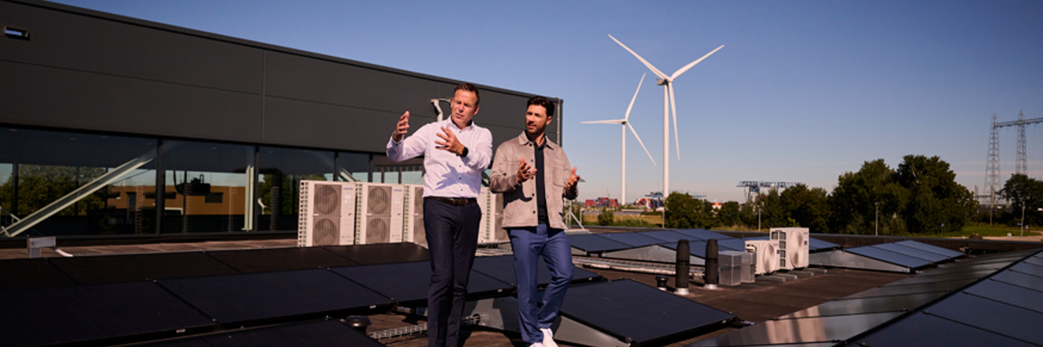 Twee mannen in gesprek op dak met zonnepanelen en met windmolens op de achtergrond